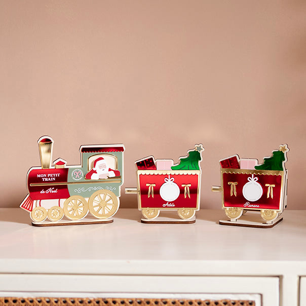 Décor en bois avec relief coloré
Représentation d’un petit train
Locomotive rouge, verte et doré avec père-noël accouché à la fenêtre
Derrière 2 wagons identiques : fond rouge avec détail doré, deux cadeaux et un sapin sorte de ceux-ci, Au centre un rond blanc, sur le coté 2 noeus dorés, et un prénom est inscrit