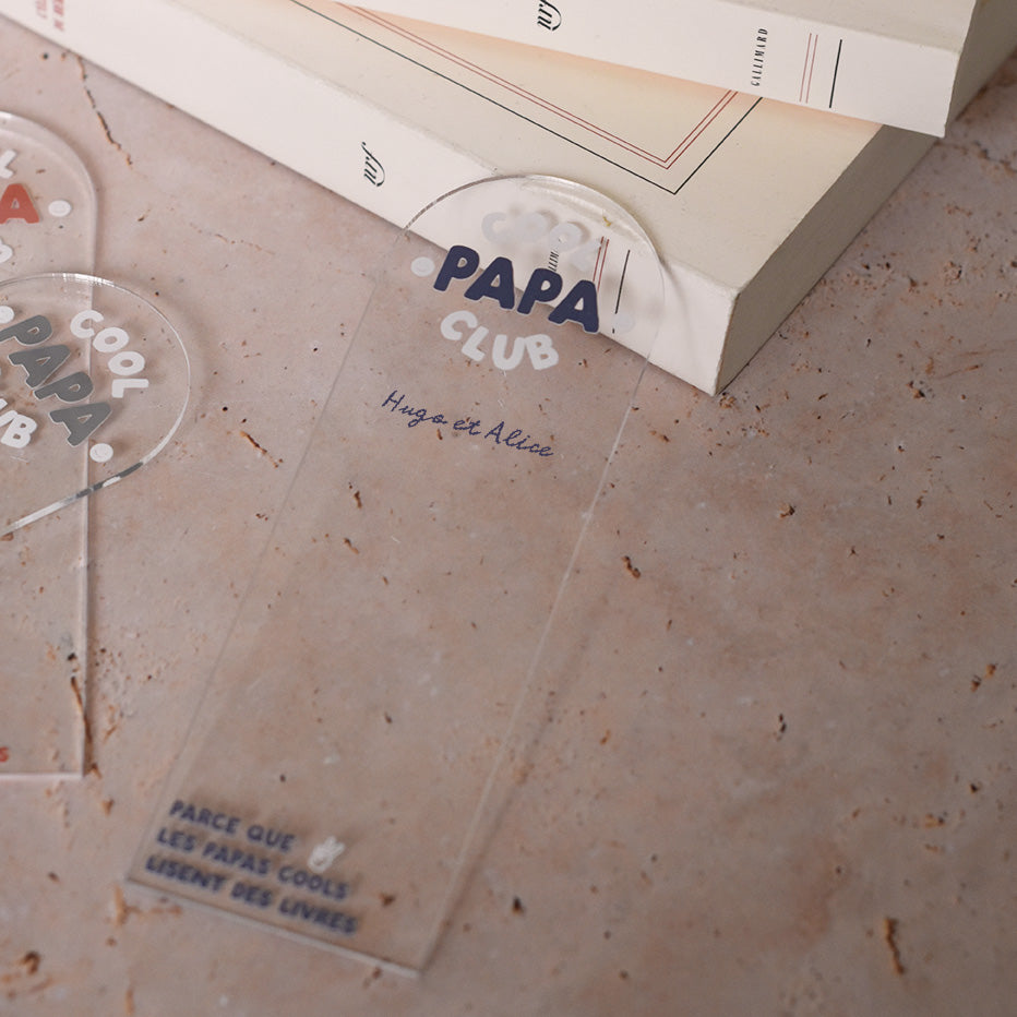 Marque-pages en plexi transparent découpe forme arche, texte blanc et bleu. Inscription dans l’arche sur 3 lignes « Cool Papa Club », 2 smiley entourent le mot « PAPA ». Les mots « cool » et « club » et les motifs sont inscrits en blanc, le reste du texte est en couleur.
En dessous est inscrit un prénoms (personnalisation du produit)
En bas du marque-page est inscrit « Parque les papas cools lisent des livres » disposé sur 3 lignes avec un dessin de main en « v »