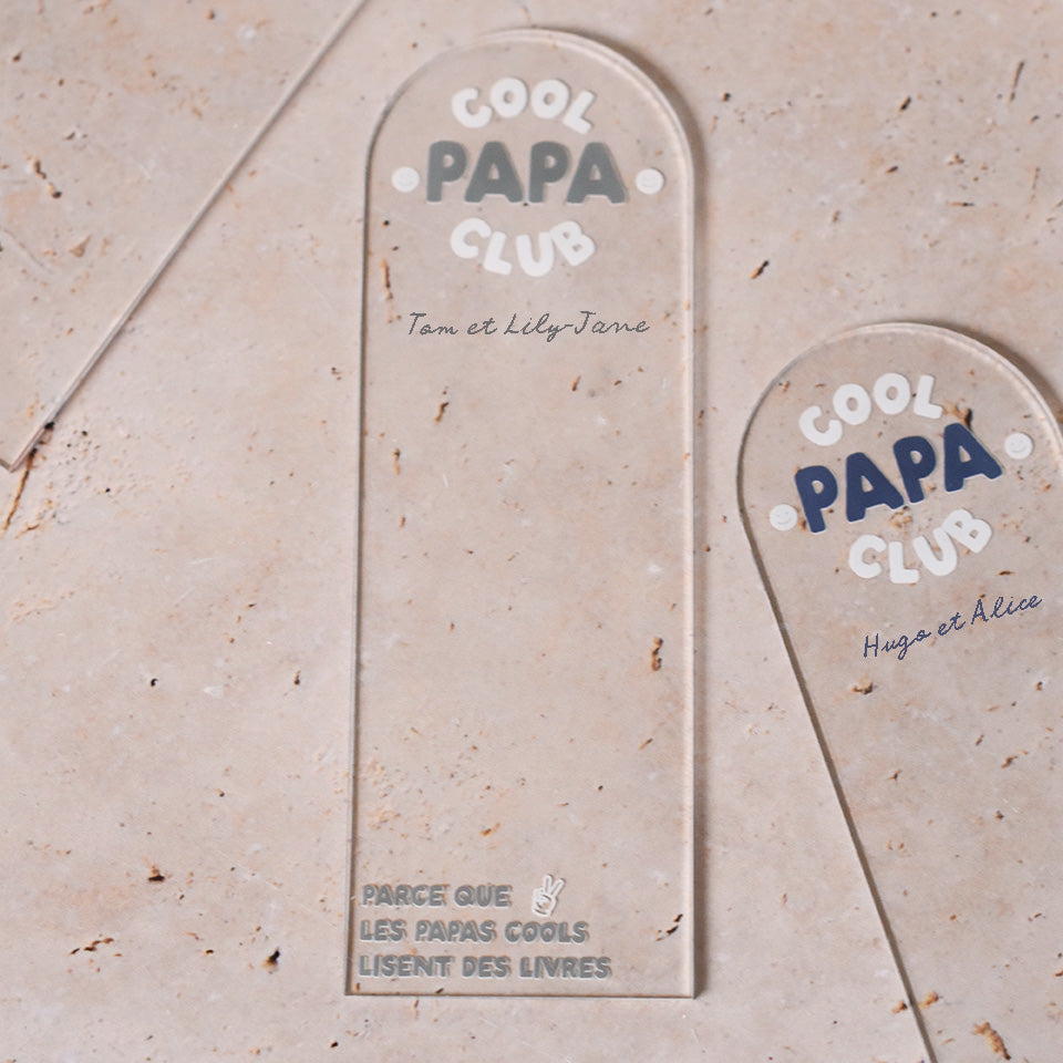 Marque-pages en plexi transparent découpe forme arche, texte blanc et coloré. Un modèle texte vert et blanc et un modèle texte bleu et blanc sont présentés.
Inscription dans l’arche sur 3 lignes « Cool Papa Club », 2 smiley entourent le mot « PAPA ».
En dessous est inscrit un prénoms (personnalisation du produit)
En bas du marque-page est inscrit « Parque les papas cools lisent des livres » disposé sur 3 lignes avec un dessin de main en « v »