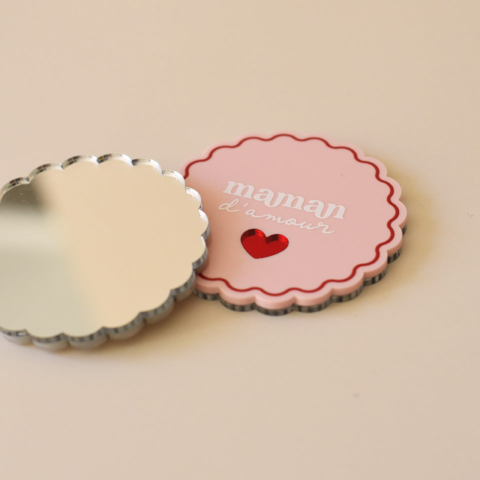 Miroir de poche forme rond festonné avec impression contour, inscription « maman d’amour » et coeur en plexi miroir incrusté au centre. Coloris face rose et rouge (fond plexi rose, contour rouge, texte blanc, coeur en plexi miroir rouge) / verso miroir argent