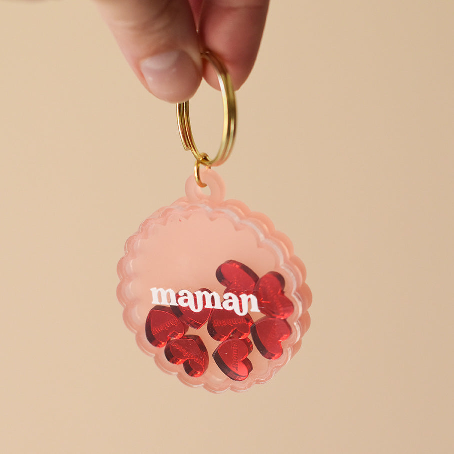 Porte-clés type en plexiglass transparent et coloré rose sur anneaux dorés. Il a une forme de cercle festonné et contient 8 mini découpes en forme de cœur rouge qui bougent librement à l’intérieur. Ces découpes sont gravées de mots associés à la maternités « tendresse, amour, bisous.. »