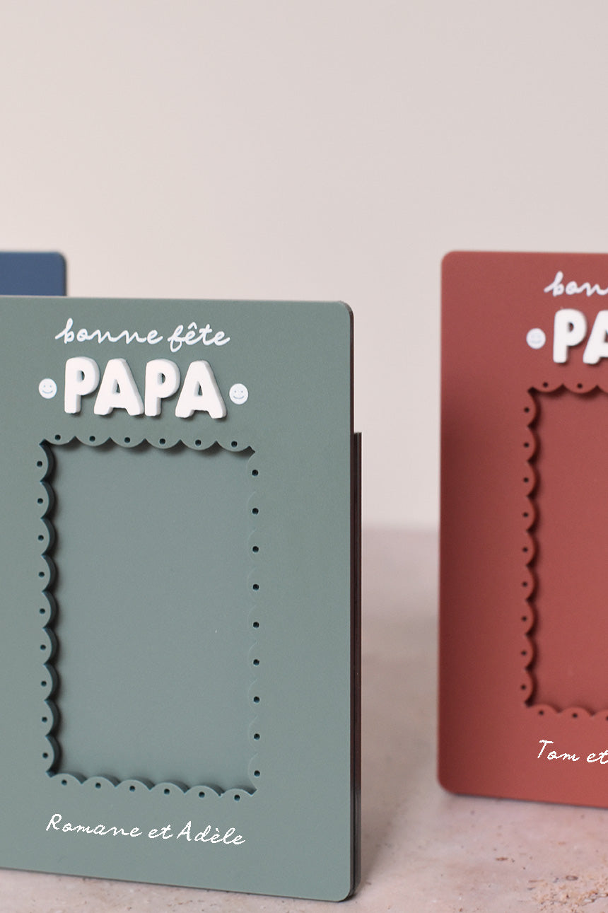 Cadre en plexi, format rectangulaire colors 3 coloris et texte blanc. Découpe de l’ouverture au centre en feston 
Inscription en haut centré sur 2 lignes, ligne 1 « bonne fête », ligne 2 « Papa » en relief plexi blanc et sur les côtés du mot 2 smileys imprimés
Inscription en bas de deux prénoms
Zoom sur le cadre vert et le relief du mot « papa »