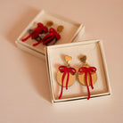 Dans une boîtes une paire de boucle d’oreilles représentant des boules de noël (décoration)
attache doré en métal et forme de coeur
découpe ronde doré et par dessus noeud rouge asymétrique