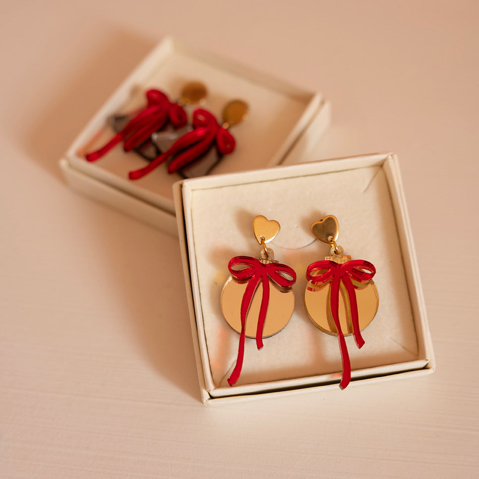 Dans une boîtes une paire de boucle d’oreilles représentant des boules de noël (décoration)
attache doré en métal et forme de coeur
découpe ronde doré et par dessus noeud rouge asymétrique