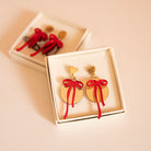 Dans une boîtes une paire de boucle d’oreilles représentant des boules de noël (décoration)
attache doré en métal et forme de coeur
découpe ronde doré et par dessus noeud rouge asymétrique