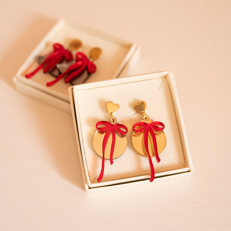 Dans une boîtes une paire de boucle d’oreilles représentant des boules de noël (décoration)
attache doré en métal et forme de coeur
découpe ronde doré et par dessus noeud rouge asymétrique