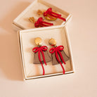 Dans une boîtes une paire de boucle d’oreilles représentant une cloche avec un noeud
attache ronde doré en métal 
cloche en colori bronze et par dessus un noeud rouge asymétrique