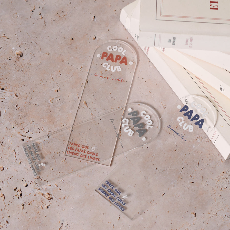 Marque-pages en plexi transparent découpe forme arche, texte blanc et coloré (3 options). Un modèle texte rouge terracotta et blanc, un modèle texte bleu et blanc et un modèle texte vert et blanc.
Inscription dans l’arche sur 3 lignes « Cool Papa Club », 2 smiley entourent le mot « PAPA ».
En dessous est inscrit un prénoms (personnalisation du produit) 
En bas du marque-page est inscrit « Parque les papas cools lisent des livres » disposé sur 3 lignes avec un dessin de main en « v » 
