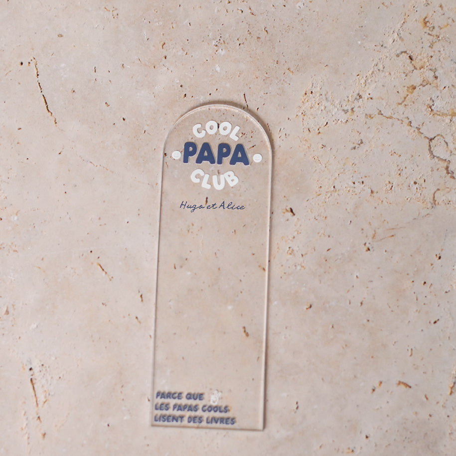 Marque-pages en plexi transparent découpe forme arche, texte blanc et bleu. Inscription dans l’arche sur 3 lignes « Cool Papa Club », 2 smiley entourent le mot « PAPA ». Les mots « cool » et « club » et les motifs sont inscrits en blanc, le reste du texte est en couleur.
En dessous est inscrit un prénoms (personnalisation du produit) 
En bas du marque-page est inscrit « Parque les papas cools lisent des livres » disposé sur 3 lignes avec un dessin de main en « v » 