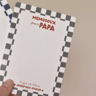 Ardoise effaçable en plexiglass blanc avec un cadre à motif damier vert, inscription centrée en haut « memodoux pour papa » couleur rouge terracotta, inscription centré en bas de deux prénoms « Hugo et Alice » et en dessous « jusqu’au étoiles » avec deux smileys sur les côtés  