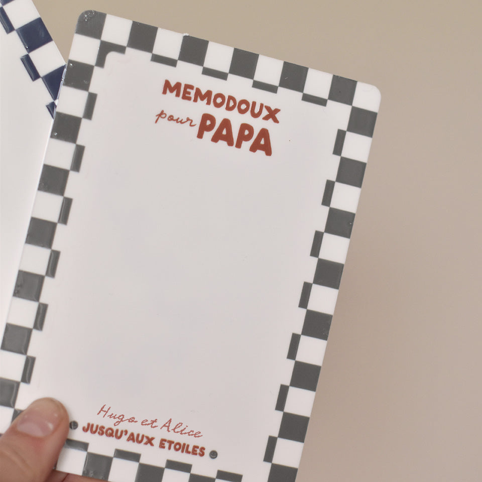 Ardoise effaçable en plexiglass blanc avec un cadre à motif damier vert, inscription centrée en haut « memodoux pour papa » couleur rouge terracotta, inscription centré en bas de deux prénoms « Hugo et Alice » et en dessous « jusqu’au étoiles » avec deux smileys sur les côtés  