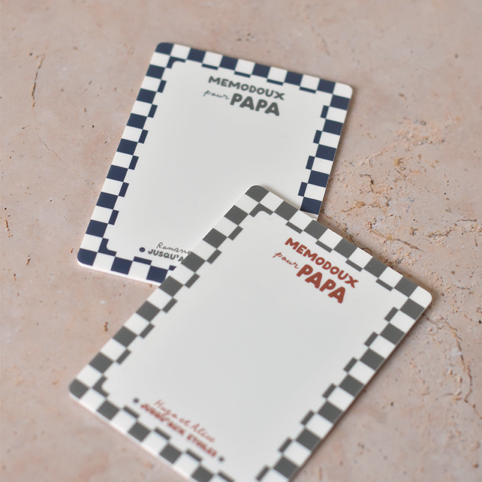 2 ardoises effaçable en plexiglass blanc avec un cadre à motif damier, inscription centrée en haut « memodoux pour papa » couleur rouge terracotta, inscription centré en bas de deux prénoms et en dessous « jusqu’au étoiles » avec deux smileys sur les côtés  
2 modèles présentés : un modèle cadre damier bleu et texte vert, un modèle cadre damier vert et texte rouge terracotta