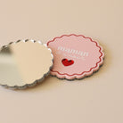 Miroir de poche forme rond festonné avec impression contour, inscription « maman d’amour » et coeur en plexi miroir incrusté au centre. Coloris face rose et rouge (fond plexi rose, contour rouge, texte blanc, coeur en plexi miroir rouge) / verso miroir argent