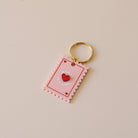 Porte-clés plexi coloré rose et rouge, design de carte à jour, forme de timbre et coeur au centre en relief, anneau doré.  Inscription maman je t’aime