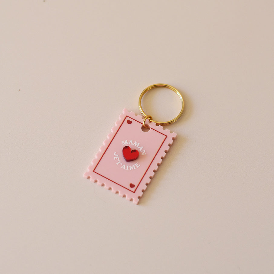 Porte-clés plexi coloré rose et rouge, design de carte à jour, forme de timbre et coeur au centre en relief, anneau doré.  Inscription maman je t’aime