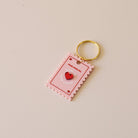 Porte-clés plexi coloré rose et rouge, design de carte à jour, forme de timbre et coeur au centre en relief, anneau doré.  Inscription reine de mon coeur 