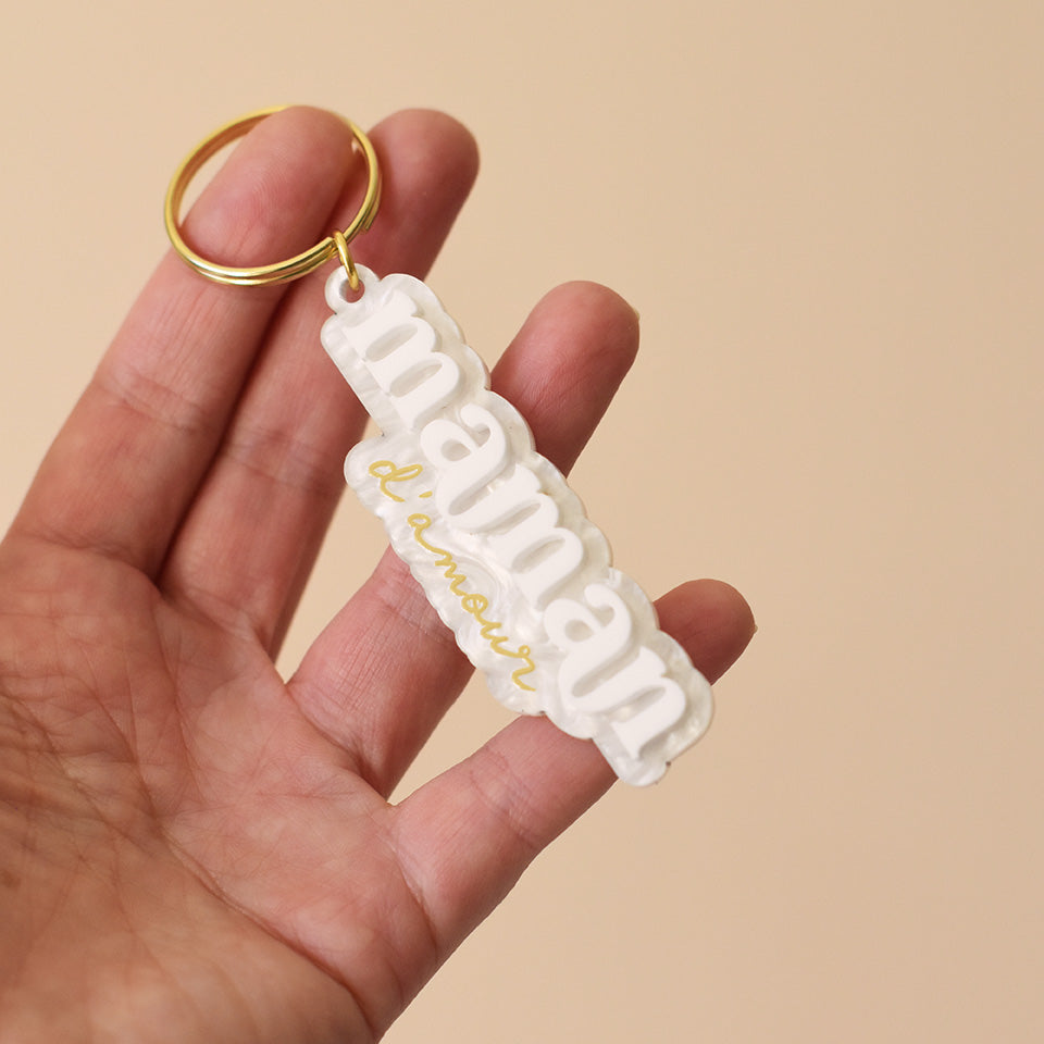 Porte-clés plexi coloré fond marbré blanc inscription « maman d’amour », « maman » en relief blanc et « d’amour » imprimé en doré, découpe à la forme du lettrage sur anneau doré