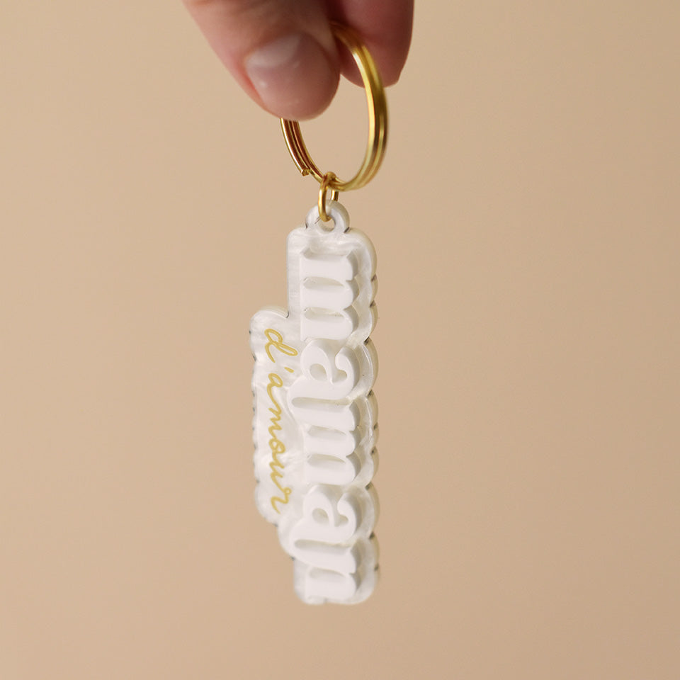 Porte-clés plexi coloré fond marbré blanc inscription « maman d’amour », « maman » en relief blanc et « d’amour » imprimé en doré, découpe à la forme du lettrage sur anneau doré